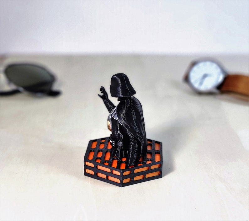May include: A black Darth Vader figurine standing on an orange and black hexagonal base. The base is designed to look like the Death Star trench run.
