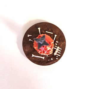 Puede incluir: Un postre de chocolate con un centro rojo y naranja, coronado con una decoraci&oacute;n gris en forma de estrella. Caramelos blancos con forma de hueso y otras decoraciones blancas est&aacute;n dispuestas alrededor del centro sobre una base marr&oacute;n oscuro texturizada.
