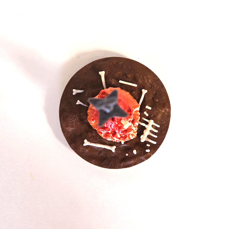 Puede incluir: Un postre de chocolate con un centro rojo y naranja, coronado con una decoraci&oacute;n gris en forma de estrella. Caramelos blancos con forma de hueso y otras decoraciones blancas est&aacute;n dispuestas alrededor del centro sobre una base marr&oacute;n oscuro texturizada.