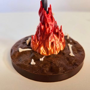 Puede incluir: Escultura impresa en 3D que representa una llama estilizada en tonos rojos y naranjas, que emerge de una base marr&oacute;n texturizada. Peque&ntilde;os elementos con forma de hueso est&aacute;n dispersos alrededor de la base.