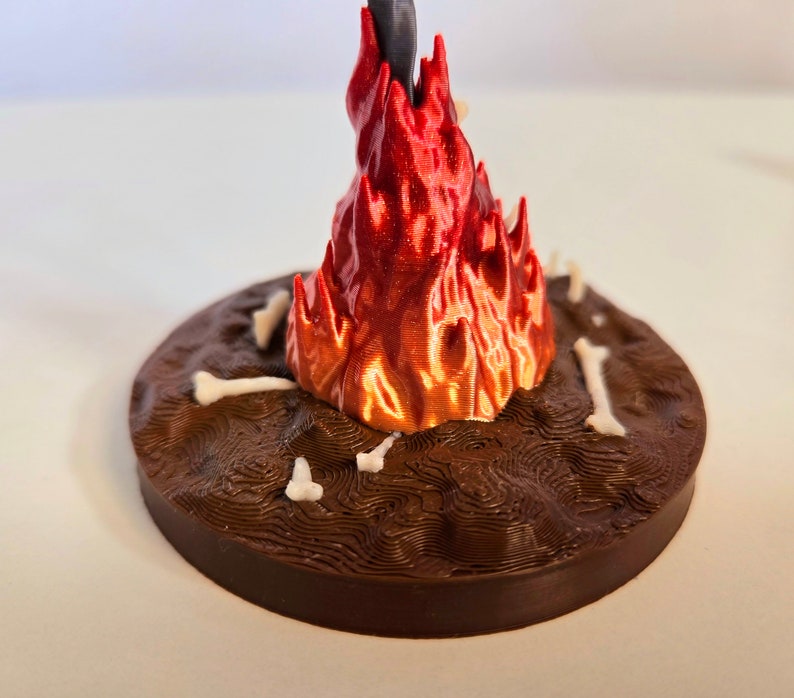 Puede incluir: Escultura impresa en 3D que representa una llama estilizada en tonos rojos y naranjas, que emerge de una base marr&oacute;n texturizada. Peque&ntilde;os elementos con forma de hueso est&aacute;n dispersos alrededor de la base.