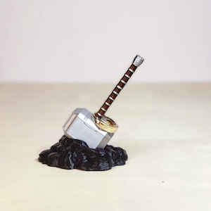 May include: A silver and brown Mjolnir ring holder with two gold rings. The holder is sitting on a black base.
