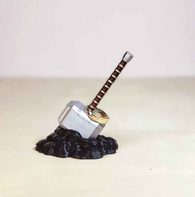 May include: A silver and brown Mjolnir ring holder with two gold rings. The holder is sitting on a black base.