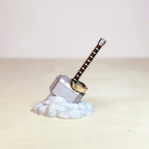 May include: A silver Mjolnir hammer ring holder with a brown handle and a gold ring on top. The holder is sitting on a white cloud-like base.