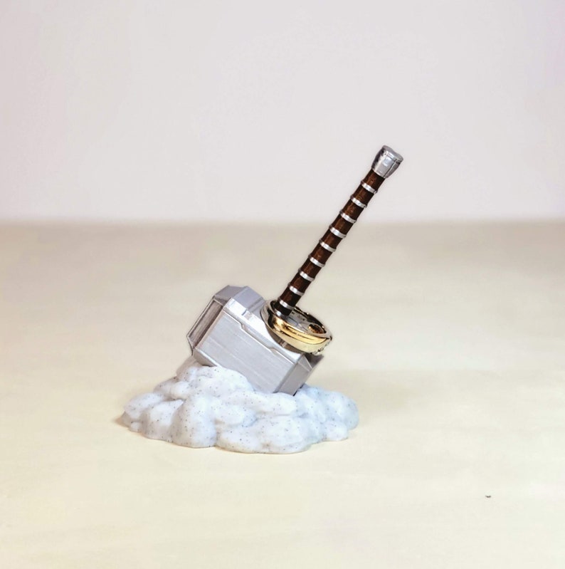May include: A silver Mjolnir hammer ring holder with a brown handle and a gold ring on top. The holder is sitting on a white cloud-like base.