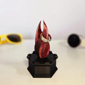 May include: A red 3D printed ring holder shaped like a flame with a black hexagonal base. A gold ring is resting on the top of the holder.