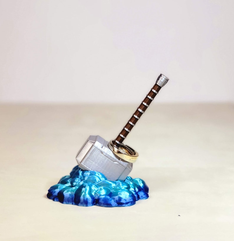 May include: A silver Mjolnir ring holder with a brown handle, sitting on a blue and teal base. Two gold rings are placed on the handle.