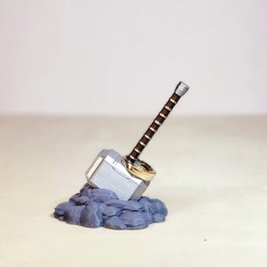 May include: A silver and brown Mjolnir hammer ring holder with two gold rings. The hammer is resting on a bed of grey rocks.