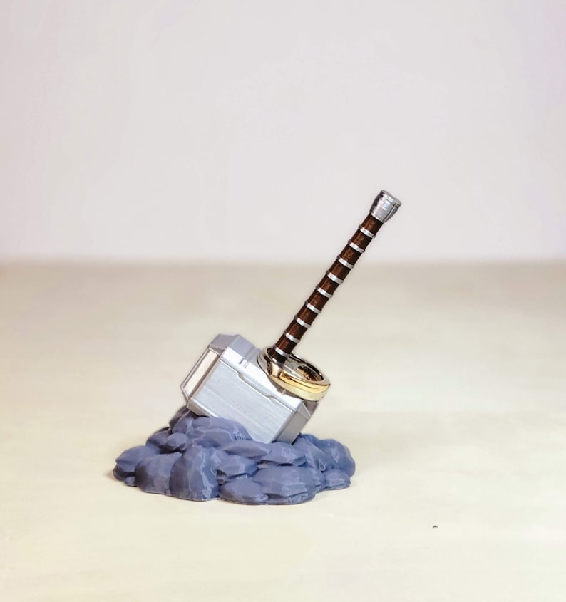 May include: A silver and brown Mjolnir hammer ring holder with two gold rings. The hammer is resting on a bed of grey rocks.