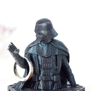 May include: A black plastic figurine of Darth Vader holding a silver ring in his outstretched hand. The figurine is on a black base.
