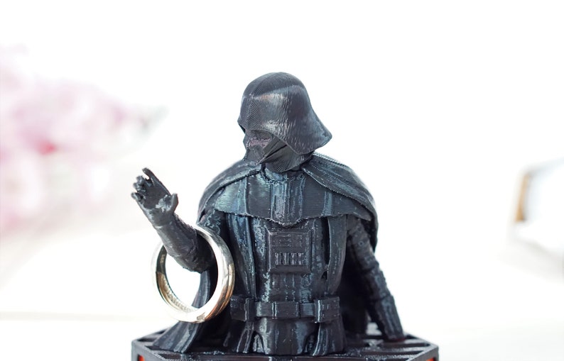 May include: A black plastic figurine of Darth Vader holding a silver ring in his outstretched hand. The figurine is on a black base.