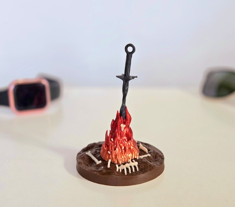 Puede incluir: Escultura en miniatura detallada de una hoguera con una espada. Las llamas son de color rojo brillante y naranja, con una espada oscura que se eleva desde el centro. La base es marr&oacute;n con peque&ntilde;os detalles &oacute;seos.