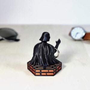 May include: A black Darth Vader figurine with a raised hand, sitting on a hexagonal orange and black platform.