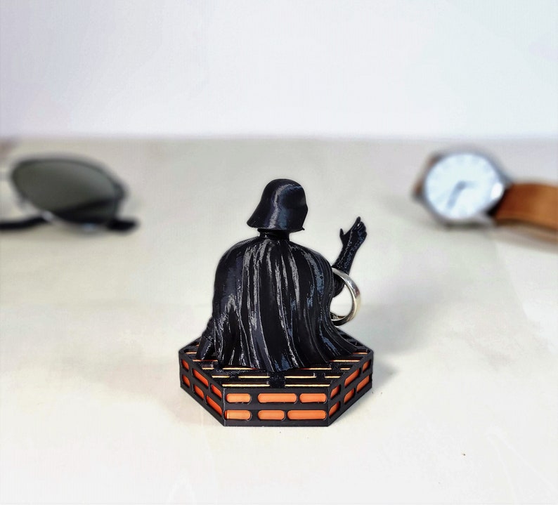 May include: A black Darth Vader figurine with a raised hand, sitting on a hexagonal orange and black platform.