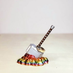 May include: A silver Mjolnir hammer ring holder with two gold rings on top, sitting on a rainbow-coloured base.