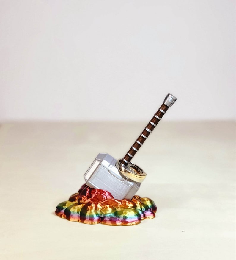 May include: A silver Mjolnir hammer ring holder with two gold rings on top, sitting on a rainbow-coloured base.