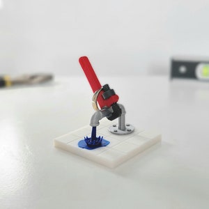 Puede incluir: Una miniatura de fontanería con una llave inglesa roja, un grifo plateado y un chorro de líquido azul. El accesorio está montado sobre una base de baldosas blancas. Un anillo dorado está unido a la llave.
