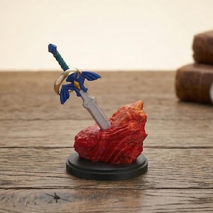 Może przedstawiać: Miniaturowa figurka miecza z niebieskim i srebrnym wzorem, osadzona w czerwonej formacji skalnej. Miecz stoi na czarnej podstawie, na drewnianej powierzchni. Inne przedmioty to zegarek i kompas.