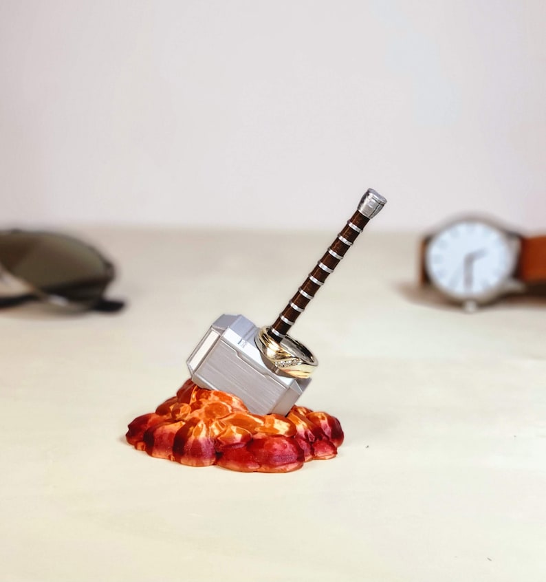 May include: A silver Mjolnir hammer ring holder with two gold rings on top, sitting on a red and orange base.