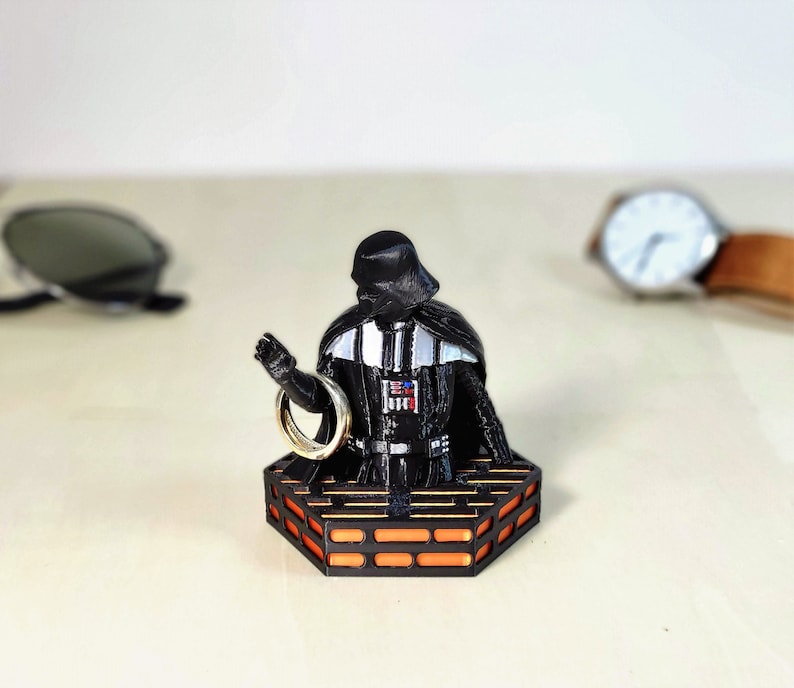 May include: A black Darth Vader figurine holding a gold ring on a black and orange platform. The platform has a hexagonal shape and resembles a Star Wars themed ring holder.