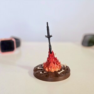 Puede incluir: Escultura en miniatura de una hoguera con una espada negra que emerge de las llamas rojas y naranjas. La base circular marr&oacute;n presenta detalles de huesos. Un objeto decorativo &uacute;nico.