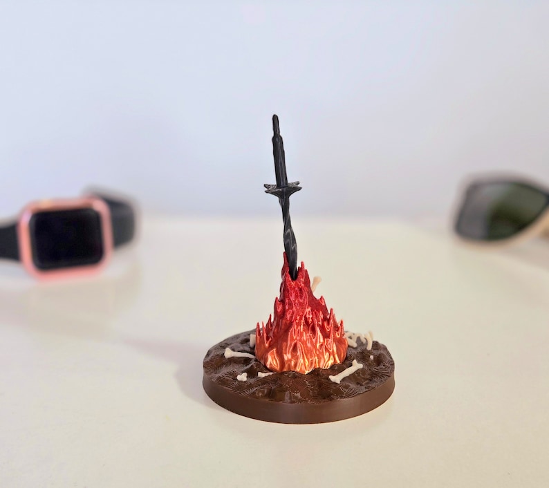 Puede incluir: Escultura en miniatura de una hoguera con una espada negra que emerge de las llamas rojas y naranjas. La base circular marr&oacute;n presenta detalles de huesos. Un objeto decorativo &uacute;nico.