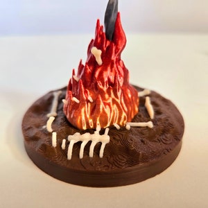 Puede incluir: Una escena de fogata en miniatura impresa en 3D. La base es una plataforma circular marr&oacute;n oscuro con una superficie texturizada. Una llama roja y naranja vibrante se eleva desde el centro, con estructuras blancas en forma de hueso alrededor de la base. Un objeto negro est&aacute; en el centro de la llama.