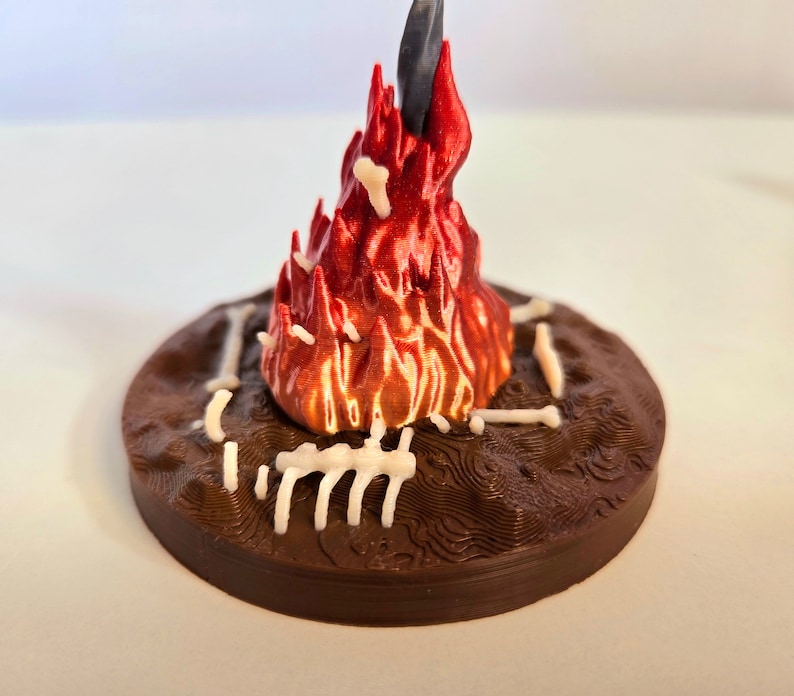 Puede incluir: Una escena de fogata en miniatura impresa en 3D. La base es una plataforma circular marr&oacute;n oscuro con una superficie texturizada. Una llama roja y naranja vibrante se eleva desde el centro, con estructuras blancas en forma de hueso alrededor de la base. Un objeto negro est&aacute; en el centro de la llama.