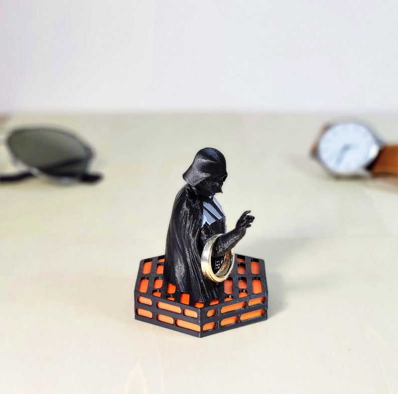 May include: A black Darth Vader figurine holding a silver ring, sitting on an orange and black brick-shaped ring holder.