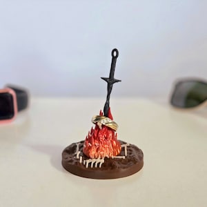Puede incluir: Una escultura en miniatura que representa una hoguera con una espada negra y dos anillos de oro. Las llamas son rojas y naranjas, y la base es marr&oacute;n. Un original soporte para anillos.