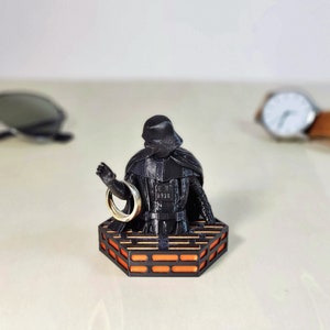 May include: A black Darth Vader figurine holding a gold ring, standing on an orange and black platform.