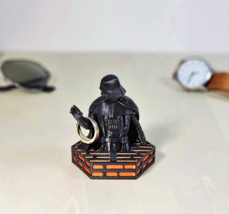 May include: A black Darth Vader figurine holding a gold ring, standing on an orange and black platform.