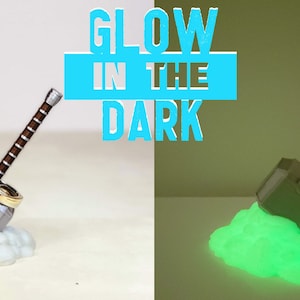 May include: A silver and brown Mjolnir hammer prop glows green in the dark. The hammer is on a white cloud-like base. The text "GLOW IN THE DARK" is in blue letters on a white background.