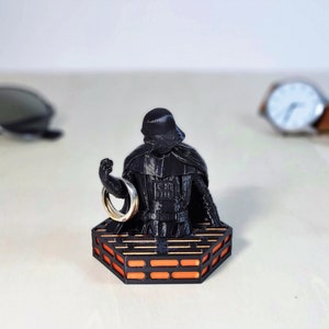 May include: A black Darth Vader figurine holding a silver ring, standing on a black and orange platform.