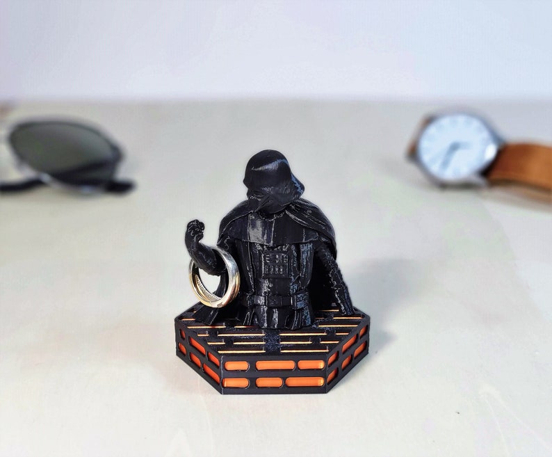 May include: A black Darth Vader figurine holding a silver ring, standing on a black and orange platform.
