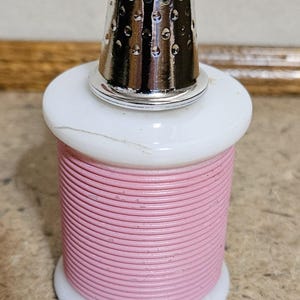 May include: A sewing-themed decorative item. It features a pink thread spool with a white base and top, topped with a silver thimble. The thimble has a perforated design. The item is likely for display or as a collectible.