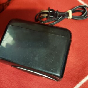 May include: A black rectangular device with a rounded top and a silver trim. A black power cord with a two-prong plug is coiled and secured with a rubber band. The device is on a red surface.