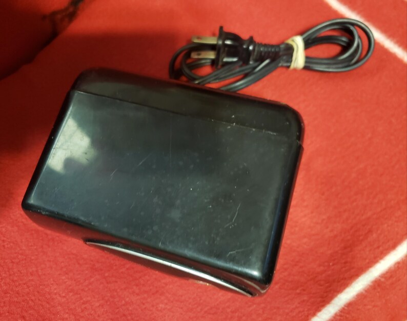 May include: A black rectangular device with a rounded top and a silver trim. A black power cord with a two-prong plug is coiled and secured with a rubber band. The device is on a red surface.