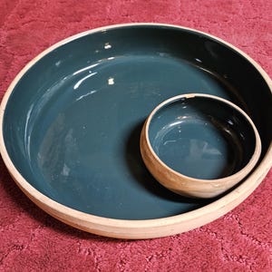 May include: Two-piece ceramic serving set. The large, round bowl has a teal interior and a natural, unglazed rim. A smaller, matching bowl sits inside. The set is on a red surface.