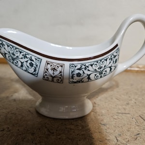 May include: White ceramic gravy boat with a decorative floral pattern in teal and brown. The gravy boat has a curved spout, a handle, and a pedestal base. The rim is trimmed with a brown band.
