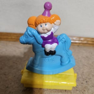 May include: A vintage Cabbage Patch Kids toy featuring a doll riding a blue rocking horse. The doll has orange hair, a purple dress, and a party hat. The base is yellow, and the toy is marked with "CPK".