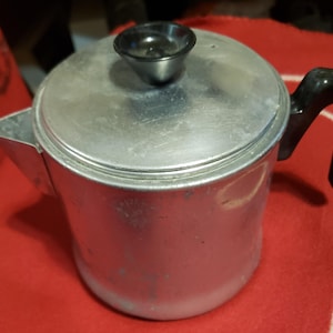 Puede incluir: Una cafetera vintage de aluminio con asa y pomo de tapa negros. La cafetera es plateada con algo de desgaste, sobre una superficie roja. La cafetera está diseñada para uso en estufa.