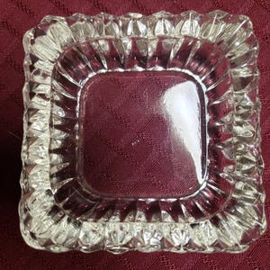 Puede incluir: Un cenicero cuadrado de vidrio transparente con un borde texturizado y acanalado. El centro del cenicero tiene una forma cuadrada sólida. El cenicero está sobre una superficie granate.