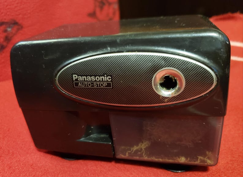 May include: Black Panasonic Auto-Stop electric pencil sharpener. The sharpener has a silver oval accent around the pencil entry point. The clear plastic shavings receptacle is visible.