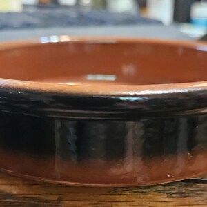May include: A round, shallow terracotta dish with a smooth, glazed finish. The interior and the upper rim feature a warm, natural clay color, while the exterior sides are coated in a dark, glossy brown glaze that transitions into the clay base.