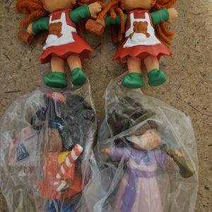 Puede incluir: Dos muñecas Cabbage Patch Kids con pelo de hilo naranja, mangas y zapatos verdes y delantales rojos y blancos. Otras dos muñecas están en envases de plástico. Una muñeca viste un atuendo rojo y blanco, y la otra un atuendo rosa y morado.