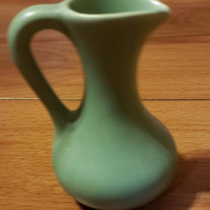 May include: A small, green ceramic pitcher with a rounded body and a curved handle. The pitcher is sitting on a wooden surface.