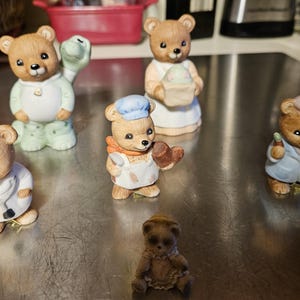 May include: A collection of small, brown bear figurines in various professions. One bear wears a doctor's coat, another a chef's hat, and another a painter's beret. Other bears are in pajamas or holding a snake. The figurines are on a reflective surface.