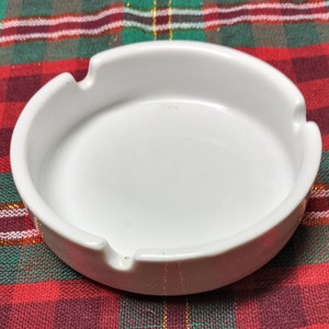 May include: A white ceramic ashtray with a round shape and a small indentation on one side. The ashtray is sitting on a red and green plaid surface.