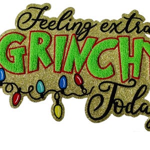 May include: A gold glittery Christmas-themed decoration with the phrase "Feeling extra GRINCHY Today" in green, red, and black. Colorful Christmas lights are also featured in the design.